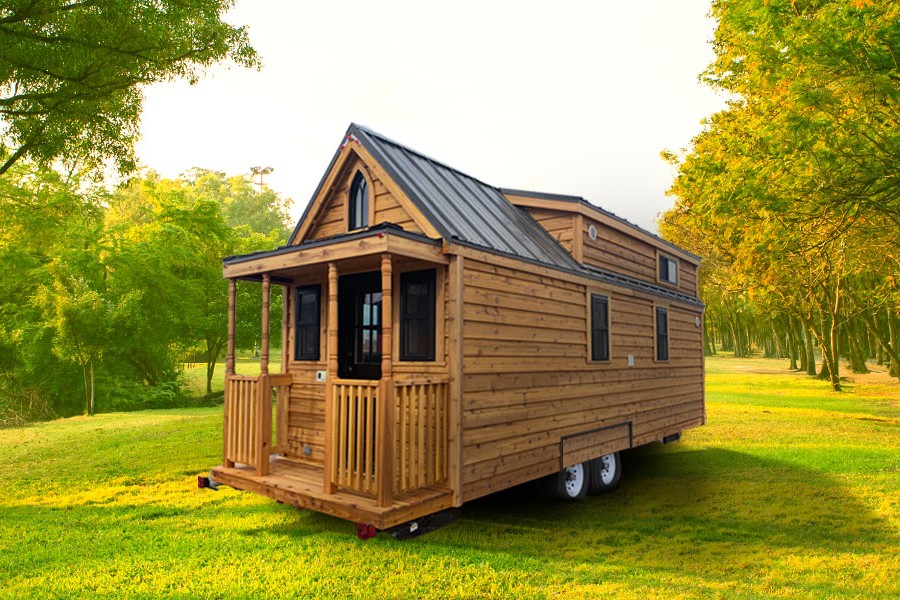 A wooden Tumbleweed tiny house with a peaked roof and front porch sits on a trailer in a sunny park surrounded by green trees and grass.