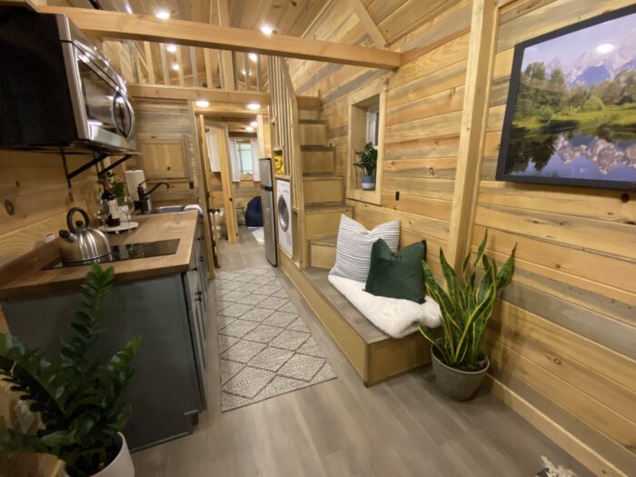 Interior of a compact wooden Tumbleweed tiny house featuring a kitchen with stainless steel appliances, a bench with cushions, a potted plant, and a view towards a small laundry area and back door.