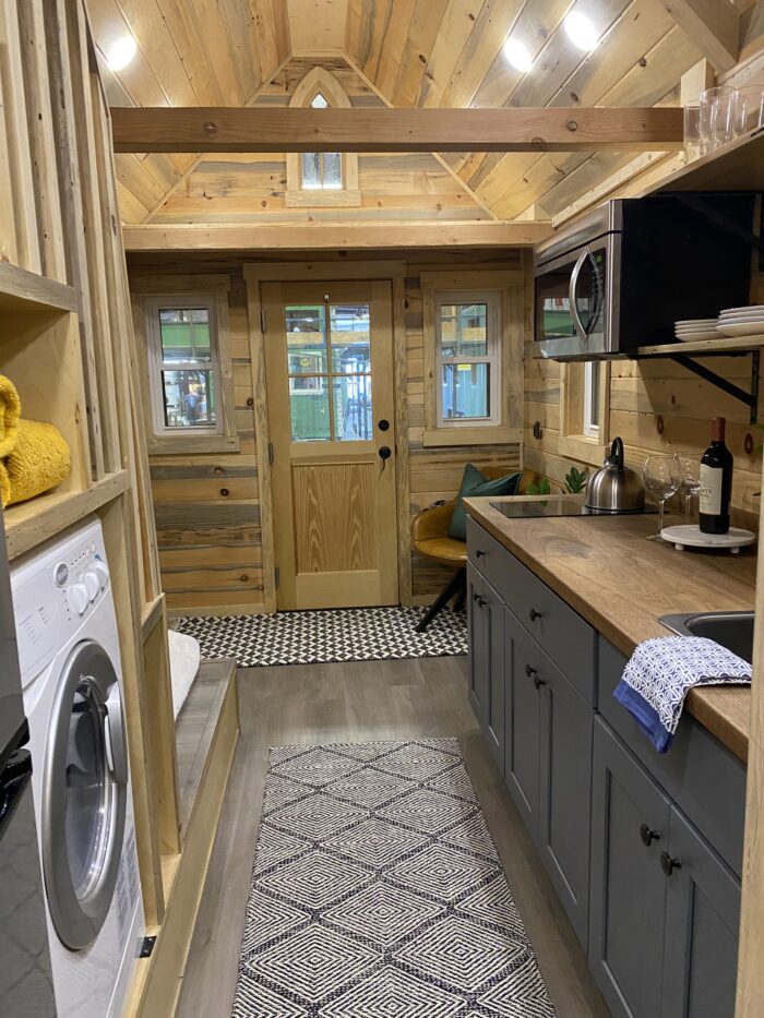 Interior of a Tumbleweed tiny house featuring a compact, wooden design with a small kitchen, front door, washing machine, and cozy seating area nestled under a loft space.