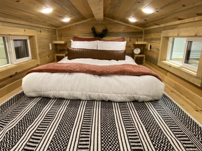 A cozy bedroom in a tiny house with a double bed, featuring white bedding and a brown accent blanket, flanked by small windows on both sides. The room has wood-paneled walls and ceiling, with a patterned rug on the floor, evoking the charm of a tumbleweed-inspired retreat.