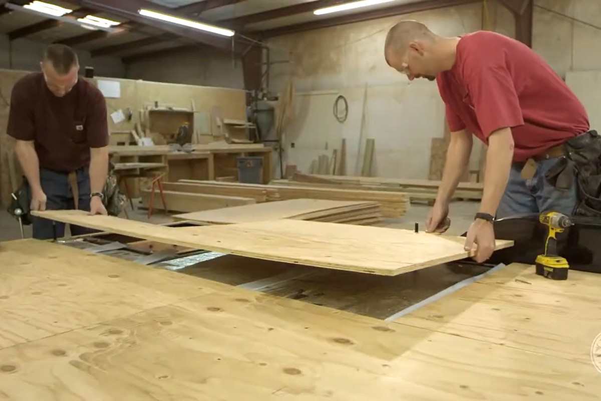 Two men in a workshop are placing a large piece of plywood on a workbench. One man is wearing a tool belt, and various tools and materials are seen in the background.