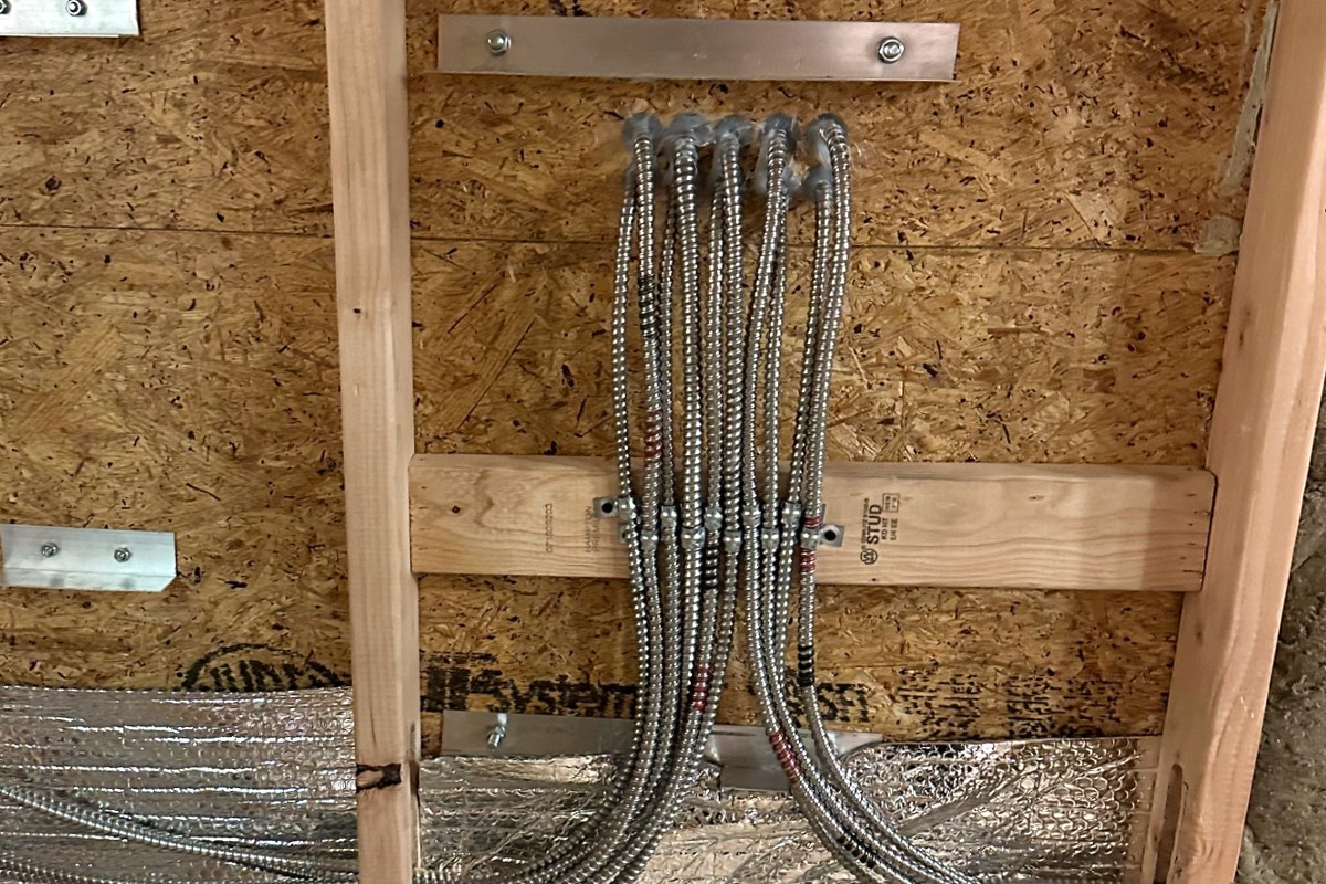 Exposed conduit wiring is installed vertically between wooden studs on an unfinished wall, secured with metal brackets and aluminum fasteners, ensuring the setup remains EMF-free.