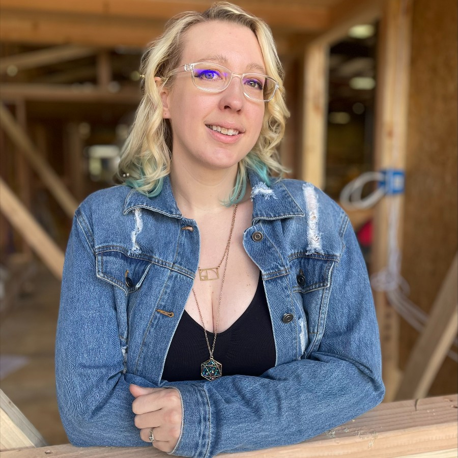 A woman with blonde hair wearing glasses and a denim jacket stands indoors, leaning on a wooden surface, reflecting her keen interest in tiny house design.