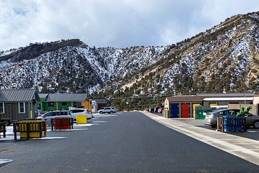 A small residential area with colorful buildings and parked cars is set against snow-dusted mountains in the background.