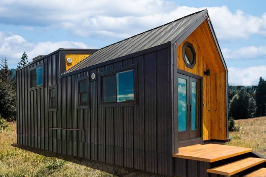 A modern black tiny house with wooden accents, large windows, and a pitched roof stands on a grassy field under a partially cloudy sky.