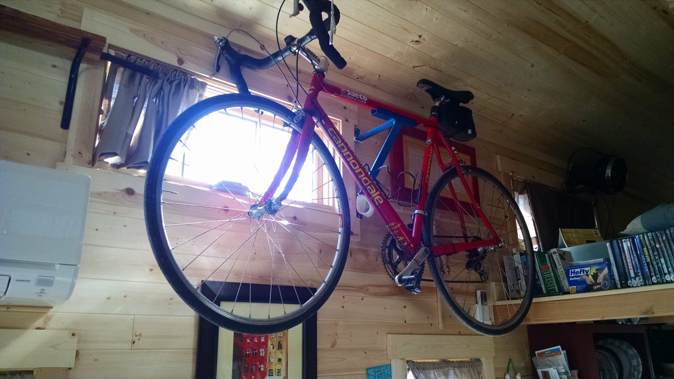 A red Cannondale road bike is mounted on a wooden wall, close to a small window in a tiny house filled with bookshelves and various household items.