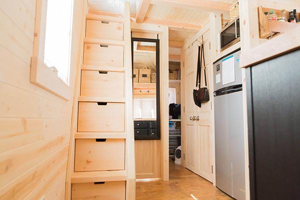 A Tumbleweed-inspired, wood-finished interior features a staircase with built-in storage, a full-length mirror, a white door, and a compact kitchen area with a refrigerator and microwave. This Tiny House maximizes space while maintaining charm and functionality.