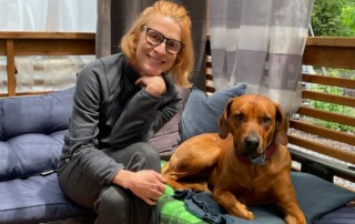 A person with glasses, mindful of their EMF sensitivity, sits on a cushioned bench next to a brown dog on a green blanket, surrounded by an outdoor setting with curtains in the background.