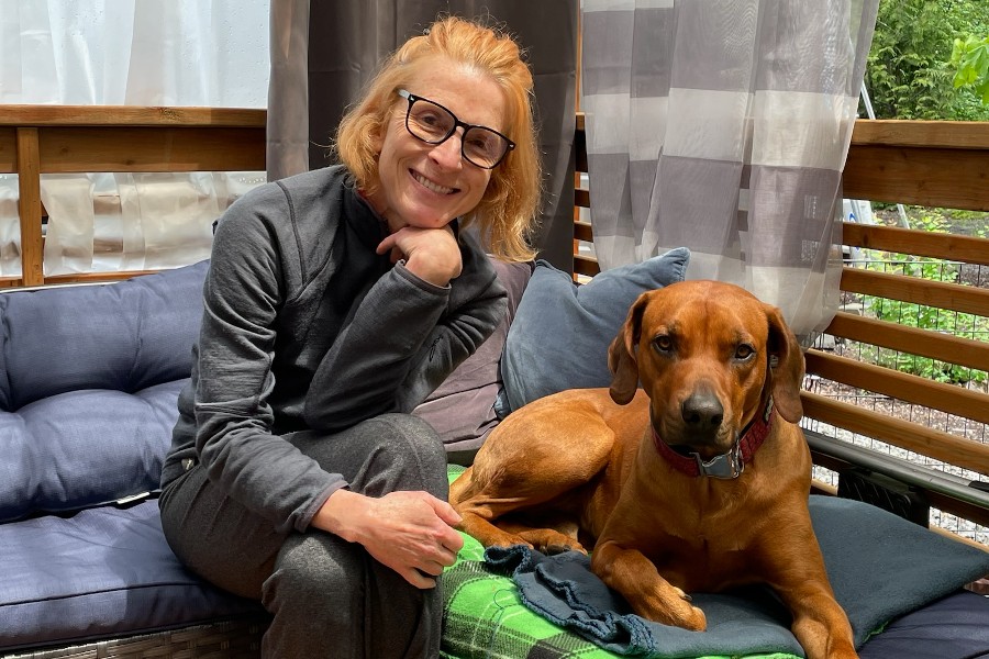 A person with glasses, mindful of their EMF sensitivity, sits on a cushioned bench next to a brown dog on a green blanket, surrounded by an outdoor setting with curtains in the background.