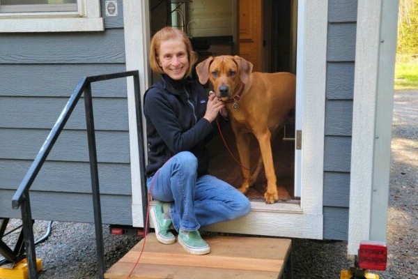 Person kneeling beside a large brown dog in the doorway of a small blue house with white trim.