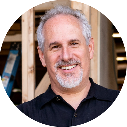 A man with gray hair and a beard smiling, standing indoors in front of a wooden construction frame and a ladder, appears to be working on his Tumbleweed tiny house.