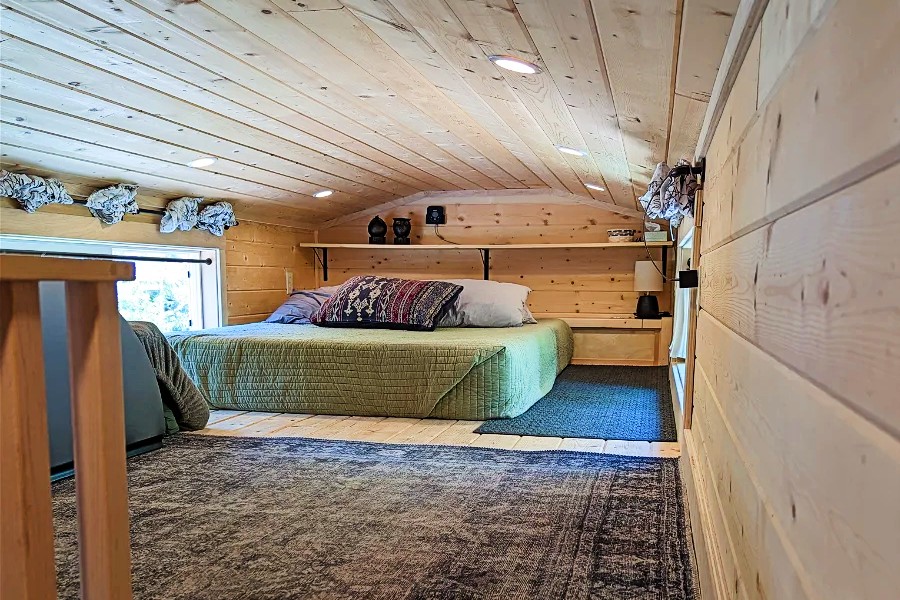 A cozy loft bedroom in this charming tiny home for sale features a wood-paneled ceiling and walls, a neatly made bed, shelves with decor, and a window adorned with ruffled curtains. Perfectly designed to embrace natural light, it's a serene retreat you'll love.