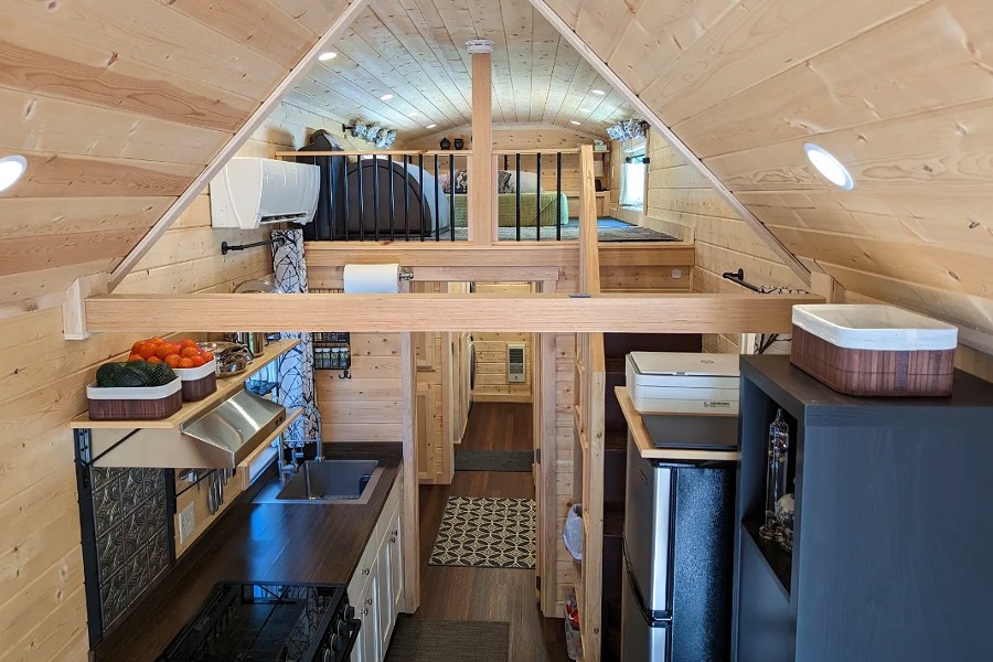 Explore the charm of this clerestory tiny home for sale, featuring a cozy interior with a loft bedroom, a sleek kitchenette complete with sink and stove, and neatly organized shelves that maximize space efficiency.