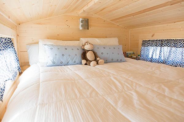 A cozy bedroom in a tiny house with wooden walls and ceiling, featuring a bed with a white comforter, two pillows, two blue patterned cushions, and a stuffed monkey toy placed against the pillows.