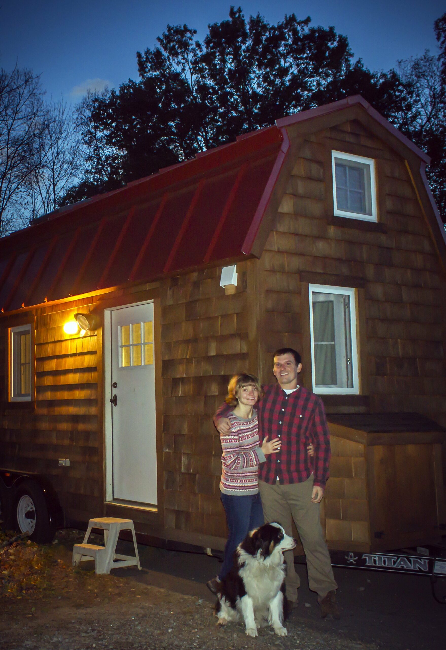 A couple stands in front of a Tumbleweed tiny house on wheels with a wooden exterior. A black and white dog sits at their feet, the scene softly illuminated by the dusk light filtering through the trees in the background.